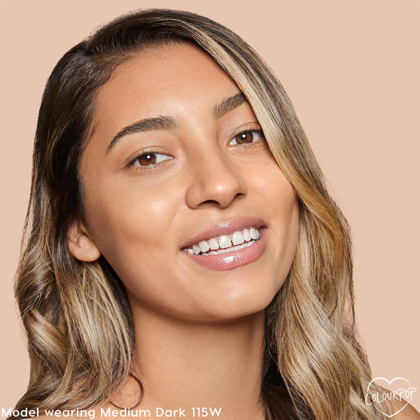 Model smiling wears ColourPop Pretty Fresh Creamy Concealer, shade Medium Dark 115 W (warm), showcasing smooth medium-dark coverage and a dewy finish on even-toned skin against a soft beige background.