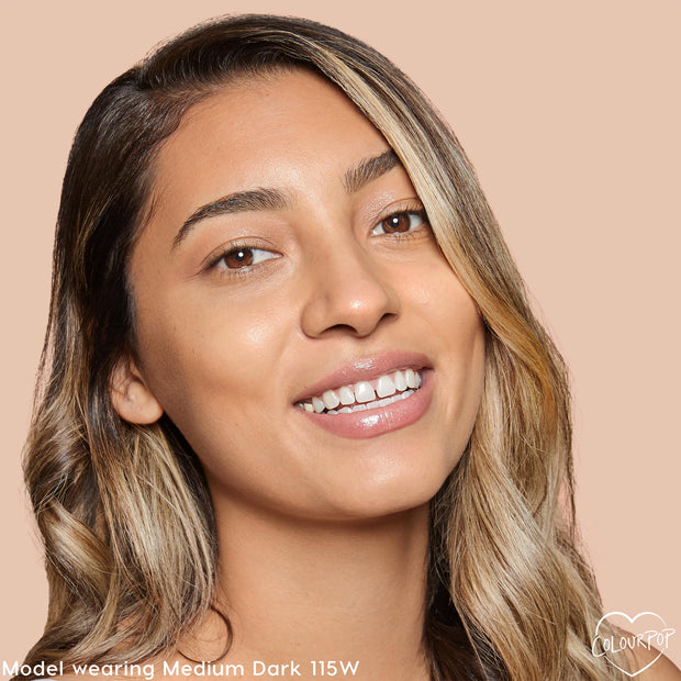 Model smiling wears ColourPop Pretty Fresh Creamy Concealer, shade Medium Dark 115 W (warm), showcasing smooth medium-dark coverage and a dewy finish on even-toned skin against a soft beige background.