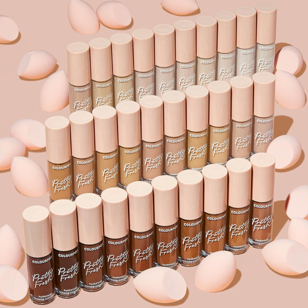 ColourPop Pretty Fresh Creamy Concealer in Fair 05 W stands in a neat grid of shades, clear tubes with blush-pink caps surrounded by soft pink makeup sponges on a nude background.