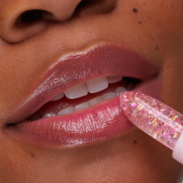 ColourPop pH Lip Balm Sing glides onto lips as a crystal-clear pink stick with iridescent flakes, delivering a glossy, rosy tint; close-up of mouth and teeth showcases hydrated shine and a subtle color-changing finish.