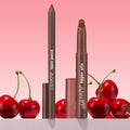 ColourPop Lippie Pencil in Pitch stands upright with cap off beside a matching Lippie Stix, both in a deep brown shade, set among glossy red cherries on a pink gradient backdrop for a bold, cherry-inspired lip look.