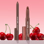 ColourPop Lippie Pencil in Pitch stands upright with cap off beside a matching Lippie Stix, both in a deep brown shade, set among glossy red cherries on a pink gradient backdrop for a bold, cherry-inspired lip look.