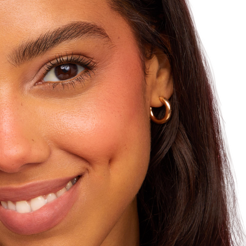 Poppi Cream Blush adding a warm peach flush to a model’s cheek, blending into natural skin for a soft, dewy finish; close-up smile with gold hoop earring on a white background, ideal to pair with ColourPop Ultra Blotted Lip.