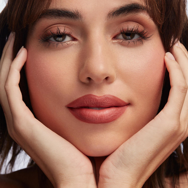 Full lips lined with Brink ColourPop Lippie Pencil, creating a warm rosy terracotta matte finish and precise shape; extreme close-up beauty shot with soft peach eyeshadow and hands gently framing the face.