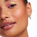 Glossy nude lips wearing Dew Drop ColourPop Lux Lip Oil, reflecting a high-shine, cushioned finish; close-up of face with warm blush, pink-and-yellow shimmer eyeshadow, and a slim gold hoop earring against a white backdrop.