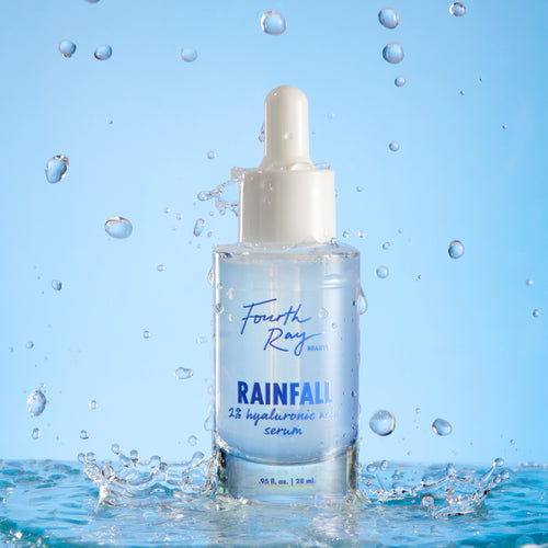 Fourth Ray Beauty Rainfall 2% HA Serum dropper bottle standing upright, catching water splashes against a light-blue backdrop, showcasing its hydrating hyaluronic acid formula; ColourPop Face Serum, 0.95 fl oz/28 mL.