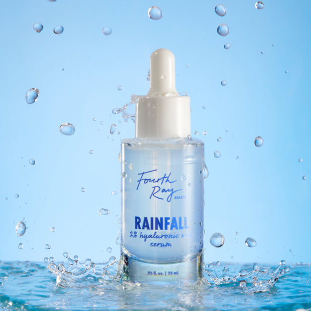 Fourth Ray Beauty Rainfall 2% HA Serum dropper bottle standing upright, catching water splashes against a light-blue backdrop, showcasing its hydrating hyaluronic acid formula; ColourPop Face Serum, 0.95 fl oz/28 mL.