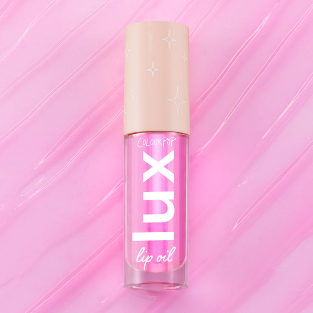 ColourPop Lux Lip Oil in Raspberry Mojito, a clear tube with bold “LUX” lettering and a beige star-print cap, displayed on a glossy swirled pink background, showcasing the vibrant pink-tinted lip oil formula.