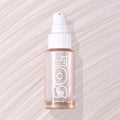 Moonstone SOL Body glow oil bottle displaying pearly pink luminizer through frosted glass with white pump, resting flat on a swirled, shimmery nude backdrop; ColourPop SOL Glow Oil for a radiant body glow.