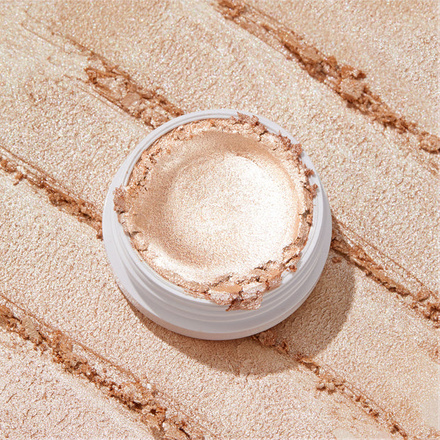 ColourPop Flexitarian Super Shock Highlighter gleaming in an open white pot, its creamy champagne‑pearl formula swatched in broad metallic streaks across the backdrop, showcasing intense shine and a smooth, blendable texture.