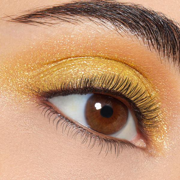 Brown eye wears ColourPop Super Shock Shadow Tankini, a metallic golden shimmer swept across the lid and crease, foiled finish with fine glitter, natural lashes and a groomed brow in a sharp studio close-up.