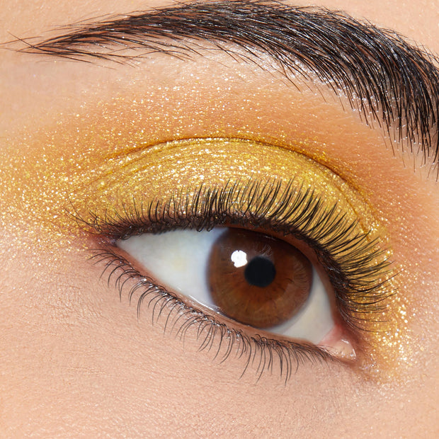 Brown eye wears ColourPop Super Shock Shadow Tankini, a metallic golden shimmer swept across the lid and crease, foiled finish with fine glitter, natural lashes and a groomed brow in a sharp studio close-up.