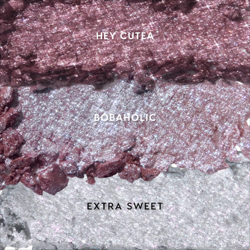 Crushed Milk Tea and Boba Super Shock Shadow swatches in three glittery shades—rose gold “Hey Cutea,” pink-lilac “Bobaholic,” and icy silver “Extra Sweet”—spread in horizontal bands on a textured background for shimmery eye makeup.