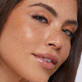 Glossy lips wearing ColourPop So Juicy Plumping Glossy Lip Oil in Lychee Jelly, plumping and high-shine; model tilts face as bronze shimmer eyeshadow and natural freckles catch studio light in a close-up beauty shot.