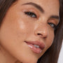 Glossy lips wearing ColourPop So Juicy Plumping Glossy Lip Oil in Lychee Jelly, plumping and high-shine; model tilts face as bronze shimmer eyeshadow and natural freckles catch studio light in a close-up beauty shot.