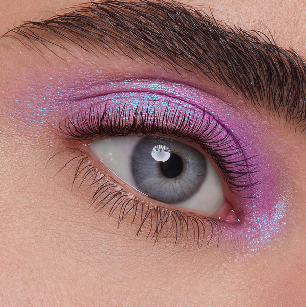 Eyelid wears ColourPop Ripple Super Shock Shadow, iridescent violet‑lavender with aqua blue sparkle, swept over lid and inner corner in an extreme close-up beauty shot highlighting lashes and brow texture.