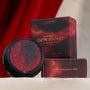 The Volturi ColourPop Super Shock Shadow showcases marbled crimson and charcoal shimmer in a black pot, catching light as it sits before Twilight: New Moon–themed boxes with gold lettering, set on a stone ledge against a rich red velvet backdrop.