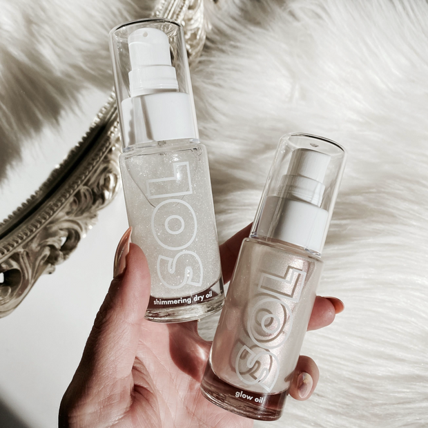Sterling Silver ColourPop SOL Shimmering Dry Oil in a clear pump bottle with fine silver shimmer, held in hand beside a matching SOL glow oil on white faux fur near an ornate mirror.