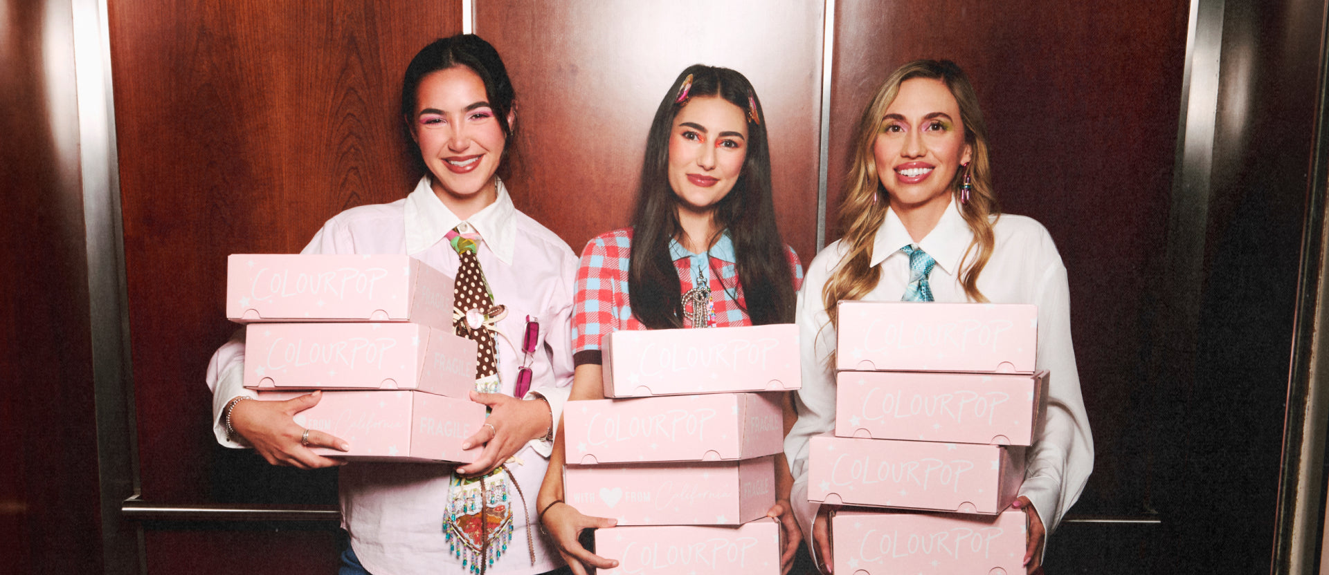 Three young women wearing colorful makeup and trendy outfits smile while holding stacks of pink ColourPop shipping boxes in a wood-paneled elevator, promoting ColourPop cosmetics delivery and fun unboxing experience in the US.