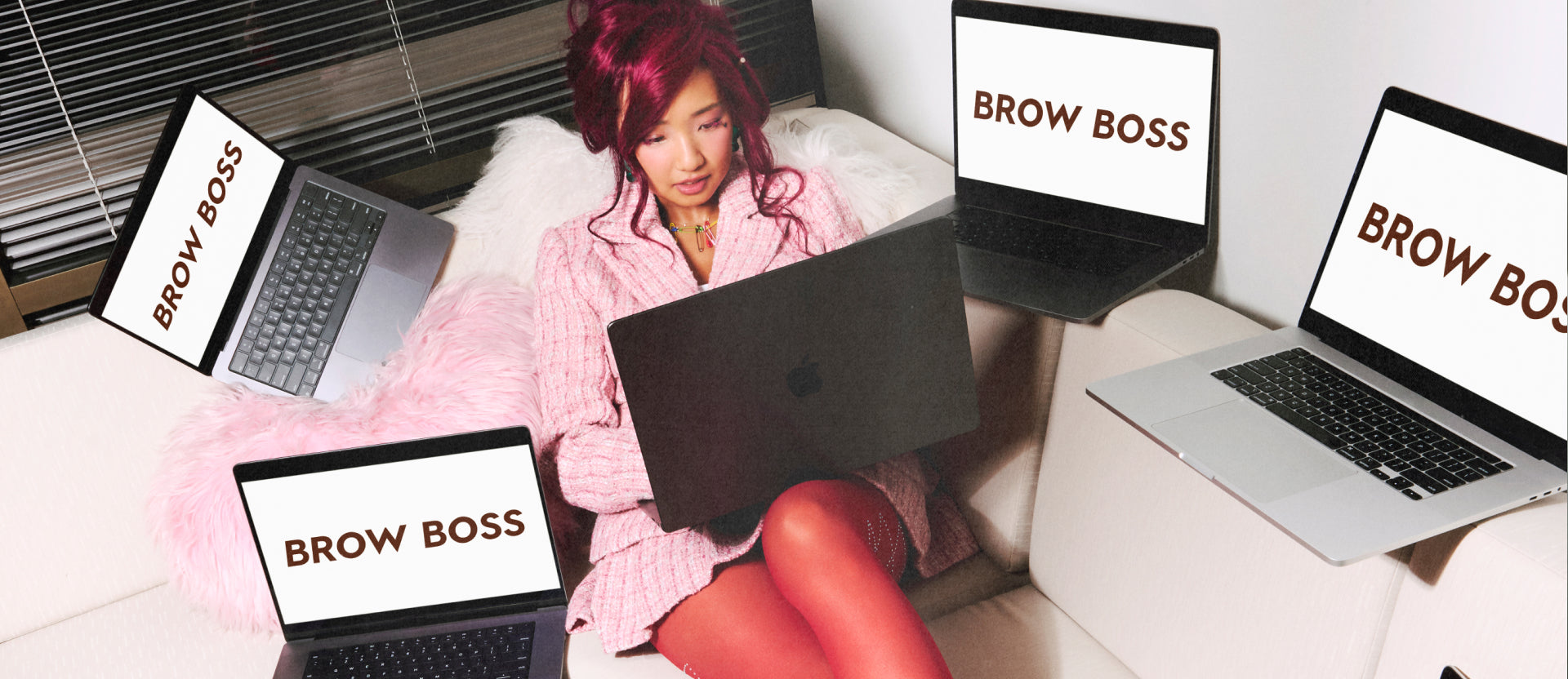 Young woman with bright pink hair and matching blazer works on a laptop while surrounded by open laptops displaying the words BROW BOSS, creating a modern, digital workspace vibe on a cozy white couch.