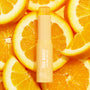 Fourth Ray Beauty Ego Boost Brightening Serum Stick with bullet twisted up, centered among vibrant orange slices on a sunny yellow background, highlighting the Best Face Scenario ColourPop Face Set.