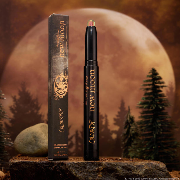 Vampire Girl ColourPop Shadow Stix stands uncapped, showing a pink‑green multichrome tip on a sleek black tube with gold lettering, its box beside it, set on a rocky surface with pine trees and a glowing full moon background.
