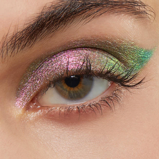 Close-up eye wearing Colourpop Galaxy Shadow Stix, showcasing metallic duochrome pink, green, and gold shimmer blended across the lid and winged outward for a bold, iridescent makeup look.