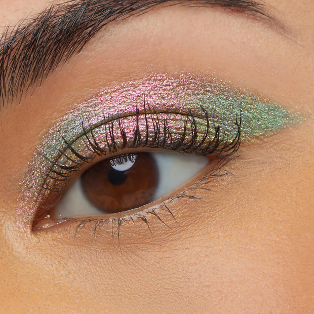 Close-up of a brown eye wearing Colourpop Galaxy Shadow Stix, showing a smooth swipe of metallic duochrome pink-to-green shimmer eyeshadow across the lid for a bold, iridescent makeup look.