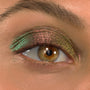 Shimmering eyelid wears Love Triangle ColourPop Shadow Stix, shifting pink‑green‑gold across the lid in a metallic multichrome finish; extreme close-up of one eye with natural lashes and brow showing color payoff.