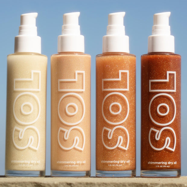 ColourPop SOL Shimmering Dry Oil in Warm Gold stands beside three deeper bronze shades, pumps up, sparkling in sunlight on a stone ledge against a blue sky; tall 3 fl oz bottles with bold SOL logo show golden shimmer.