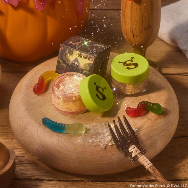 ColourPop Glitter Gel Ogres are Like Onions features an open jar of gold iridescent glitter gel with a lime green lid stamped with an ogre ear, beside a closed pot and Shrek x ColourPop box on a wooden board with gummy worms and scattered sparkles.