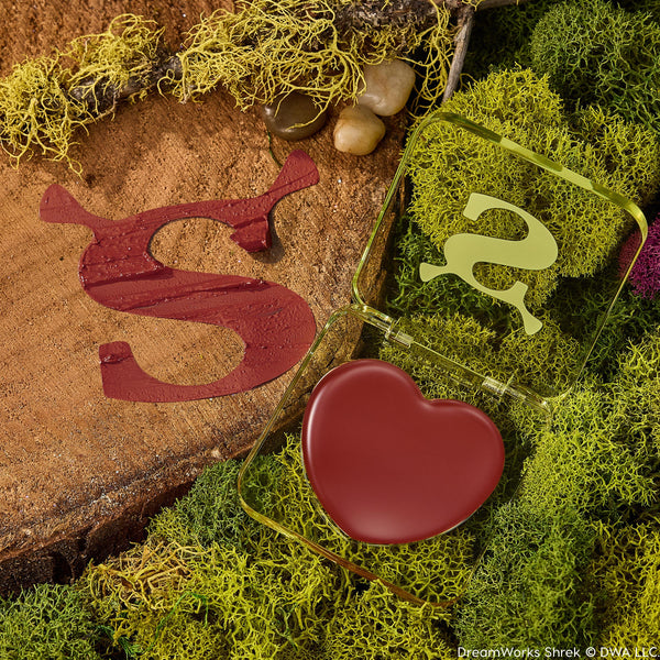 Fairytale Things ColourPop Lip and Cheek Balm sits open, revealing a heart-shaped pan of warm brick-red pigment in a translucent green, Shrek-inspired compact with an ogre-ear S, resting on moss and a wood slice beside a glossy burgundy S swatch.
