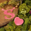 Heart-shaped pink balm in a translucent green compact sits open on moss and a wood slab with a Shrek-style S, showcasing ColourPop Lip and Cheek Balm, True Love’s Form, a lip and cheek tint in a playful swamp-inspired setting.