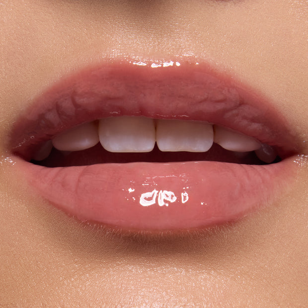 Glossy, close-up lips wearing ColourPop Ultra Glossy Lip in Totally Swamped, a sheer pink-nude shade, catching light with a glassy finish; teeth softly visible in an extreme macro beauty shot against natural skin.