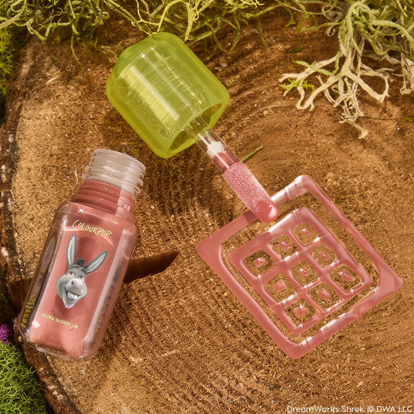 ColourPop Ultra Glossy Lip Noble Steed opens to reveal a doe-foot applicator coated in rosy mauve shimmer, with a neon-green cap and Donkey graphic bottle resting on a wood slice with moss - Shrek collab lip gloss for a high-shine finish.