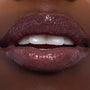 High-shine close-up of lips wearing ColourPop Ultra Glossy Lip in Ogress, a deep plum micro-shimmer gloss, reflecting light and showing a smooth, glassy finish on a model’s lips under studio lighting.