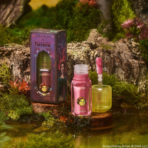 ColourPop Ultra Glossy Lip in Ogress shown with an open sparkly pink gloss tube and doe-foot applicator beside the Shrek x ColourPop illustrated box, styled on mossy swamp scenery for a whimsical look.