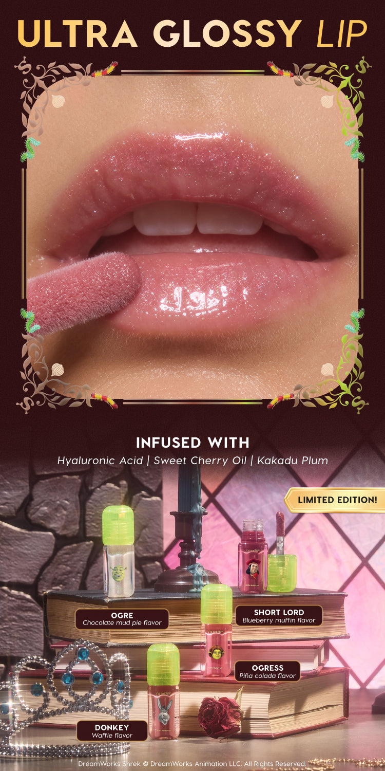 Close-up glossy pink lips being coated with Ultra Glossy Lip gloss, above a fantasy vanity scene displaying limited edition Shrek-themed Ultra Glossy Lip flavors—Ogre chocolate mud pie, Short Lord blueberry muffin, Ogress piña colada, and Donkey waffle—on stacked books with a tiara and rose decor.