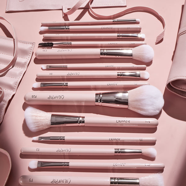 Smoke 'N Roses Brush Roll displaying a ColourPop Makeup Brush Kit: blush pink handled face and eye brushes - powder, blush, blending, liner, and spoolie - with white bristles and chrome ferrules, laid beside a soft pink roll-up case on a matching backdrop.