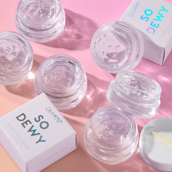 ColourPop Face & Eye Gloss in In The Clear glistening in open glass pots, clear gel catching light, arranged on a pink backdrop beside So Dewy boxes and lids, showcasing the transparent, dewy finish.