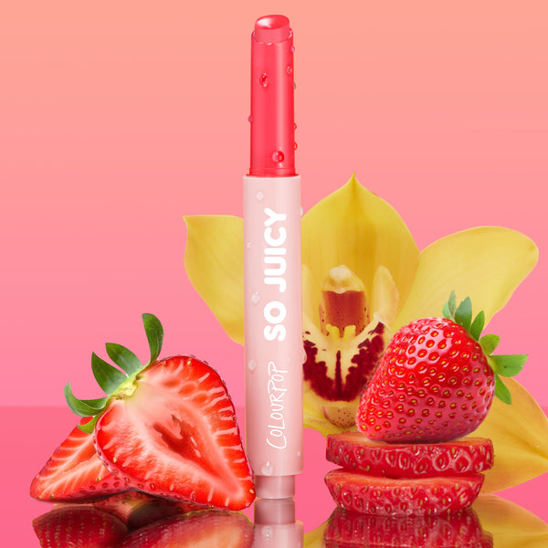Delulu shade of ColourPop So Juicy Plumping Gloss Balm with Peptides stands uncapped, showing a glossy pink bullet with dewy droplets, surrounded by sliced strawberries and a yellow orchid on a reflective surface against a pink gradient.