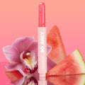 Passenger Princess shade of the ColourPop So Juicy Plumping Gloss Balm with Peptides stands upright with dewy droplets, bullet slightly twisted up, set against a pink gradient with an orchid and watermelon slices on a reflective surface.