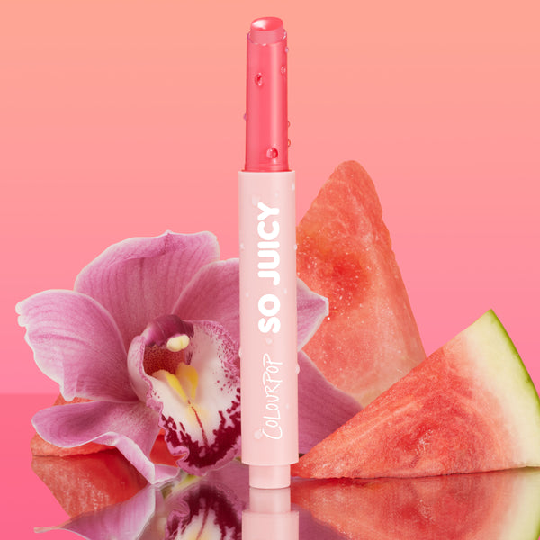 Passenger Princess shade of the ColourPop So Juicy Plumping Gloss Balm with Peptides stands upright with dewy droplets, bullet slightly twisted up, set against a pink gradient with an orchid and watermelon slices on a reflective surface.