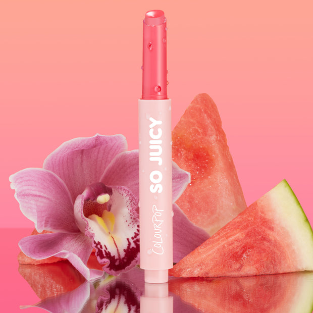 Passenger Princess shade of the ColourPop So Juicy Plumping Gloss Balm with Peptides stands upright with dewy droplets, bullet slightly twisted up, set against a pink gradient with an orchid and watermelon slices on a reflective surface.