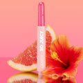Tea Sesh, the ColourPop So Juicy Plumping Gloss Balm with Peptides, stands upright with dewy droplets on its pink tube and glossy bullet exposed, flanked by a grapefruit wedge and red hibiscus on a pink gradient, reflective surface.
