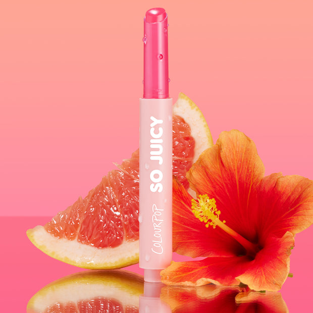 Tea Sesh, the ColourPop So Juicy Plumping Gloss Balm with Peptides, stands upright with dewy droplets on its pink tube and glossy bullet exposed, flanked by a grapefruit wedge and red hibiscus on a pink gradient, reflective surface.
