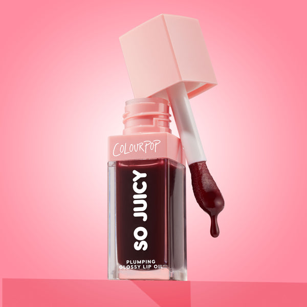ColourPop So Juicy Plumping Glossy Lip Oil in Beverly Drive stands open with the applicator lifted and a drop of deep cherry oil forming, the square clear tube and blush-pink cap set against a glossy pink gradient background.