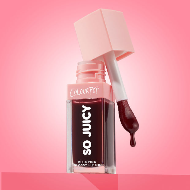 ColourPop So Juicy Plumping Glossy Lip Oil in Beverly Drive stands open with the applicator lifted and a drop of deep cherry oil forming, the square clear tube and blush-pink cap set against a glossy pink gradient background.