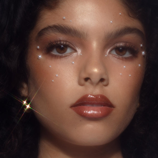 High-shine lips wearing Angel Baby shade show the ColourPop So Juicy Plumping Gloss Balm with Peptides in action, glazing a warm caramel-brown finish on a model's full face with dewy skin, shimmery eyeshadow, and pearl accents in soft studio lighting.