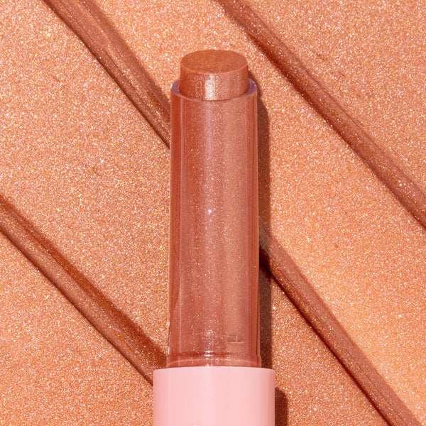 Angel Baby ColourPop So Juicy Plumping Gloss Balm with Peptides extends from a pink tube, the shimmery peach-nude bullet catching light, shown atop matching metallic swatches to highlight its glossy, plumping finish.